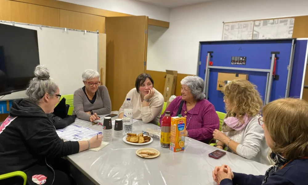 A la foto es veu un grup de gent entorn a una taula que parlen animadament entre elles sobre els temes proposats de la reunió. Sobre la taula hi ha berenar: galetes, plumcake, suc de fruita, cacaolat, aigua.