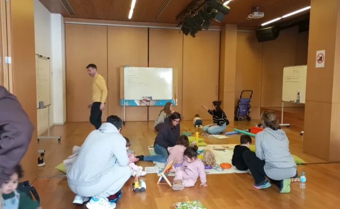 La imagen muestra diferentes familias, padres y madres con niños, en una sala polivalente de parquet, jugando en el suelo con diferentes juegos, sobre alfombras blandas. Los niños son de edades comprendidas entre los 0 y los 3 años. Hay unas pizarras con ideas de actividades anotadas.